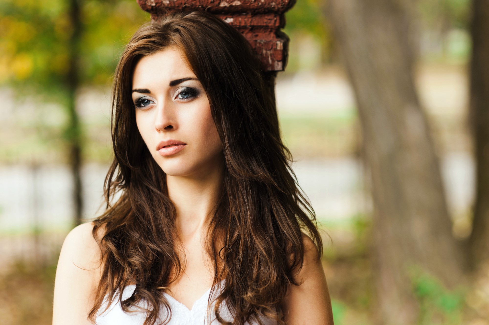 Woman with long brown hair and a contemplative expression standing outdoors, leaning against a brick pillar. She is wearing a white top, and the background is blurred with trees and greenery.