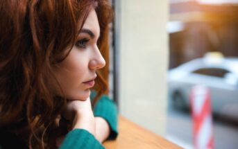 Woman with long, curly red hair gazing out of a window, resting her chin on her hands. She wears a green sweater and looks thoughtful, with blurred street and car visible outside.