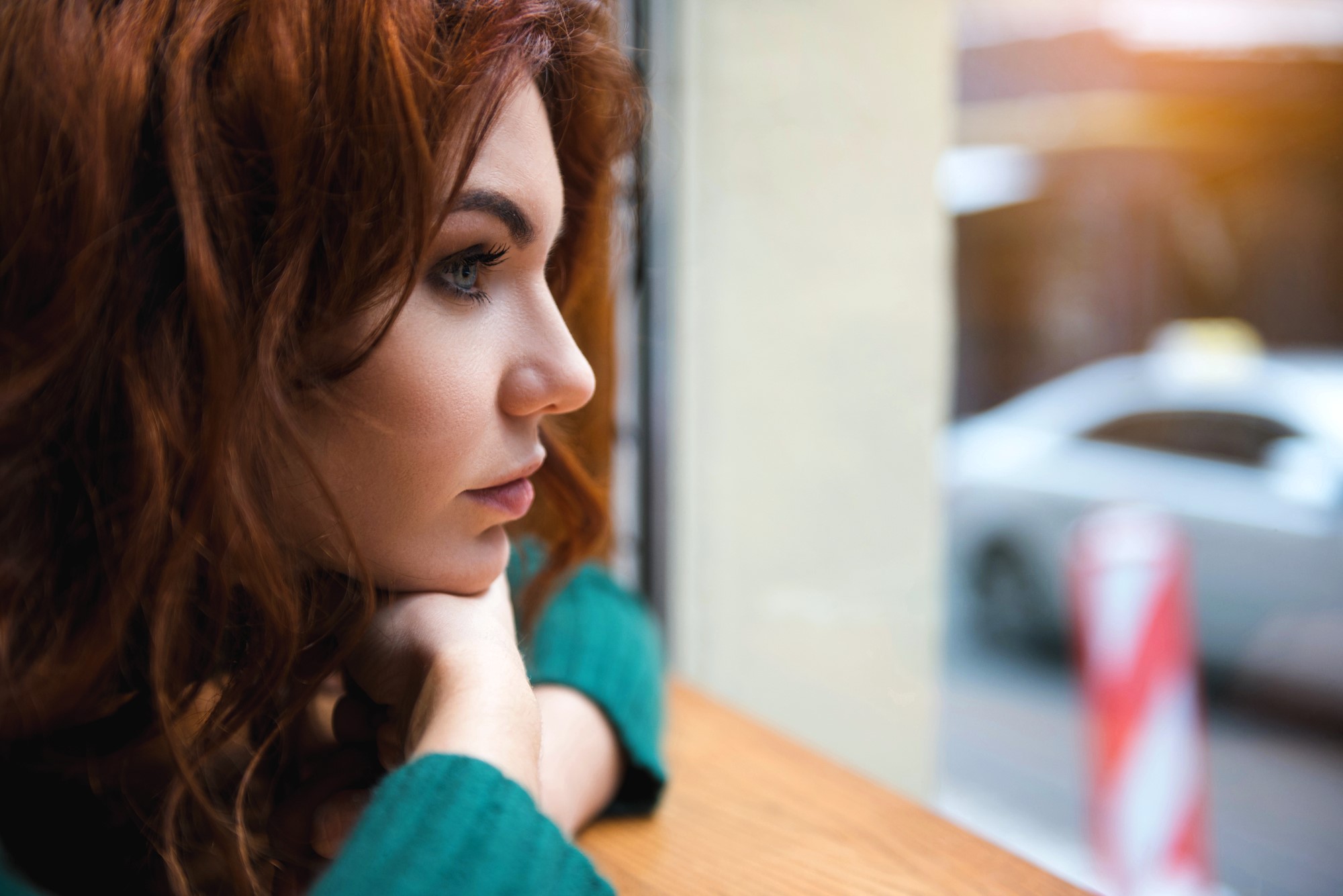 Woman with long, curly red hair gazing out of a window, resting her chin on her hands. She wears a green sweater and looks thoughtful, with blurred street and car visible outside.