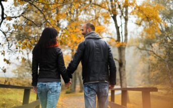 A couple walks hand-in-hand across a wooden bridge in a park during autumn. They wear black leather jackets and jeans. The trees around them are covered in vibrant yellow leaves, and the atmosphere is serene and romantic.