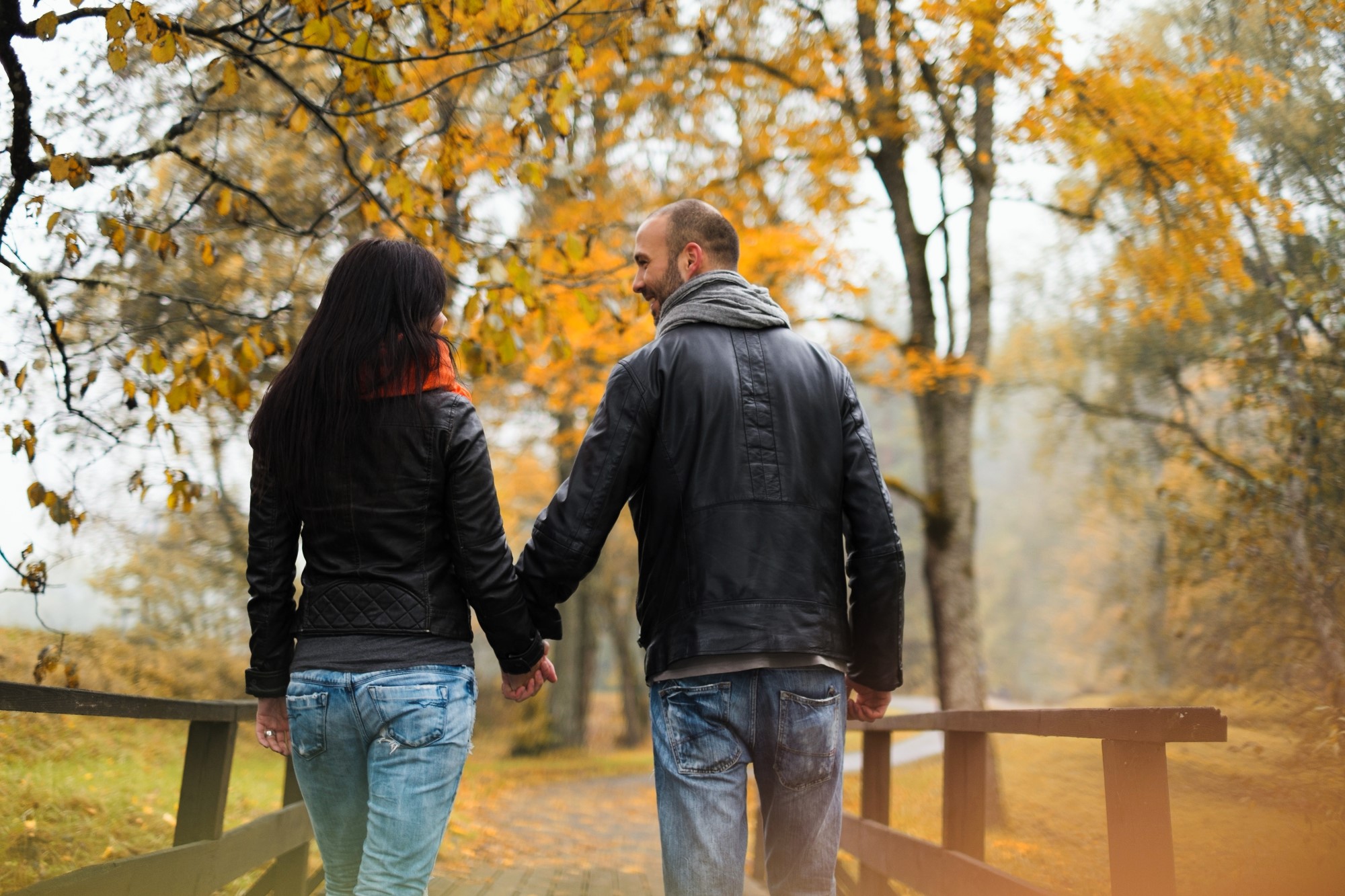 A couple walks hand-in-hand across a wooden bridge in a park during autumn. They wear black leather jackets and jeans. The trees around them are covered in vibrant yellow leaves, and the atmosphere is serene and romantic.