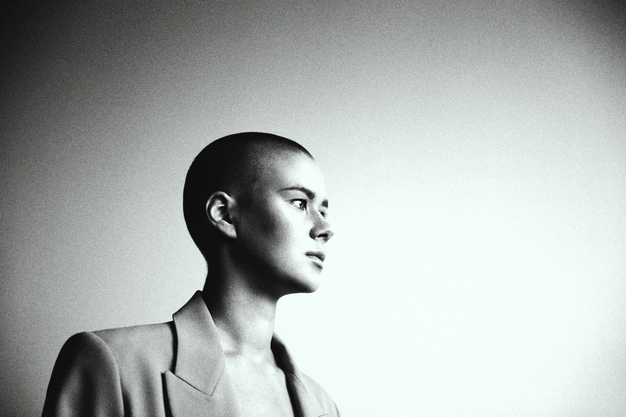 Black and white photo of a person with a shaved head, wearing a blazer. They are gazing to the right against a plain backdrop, creating a serene and contemplative mood.