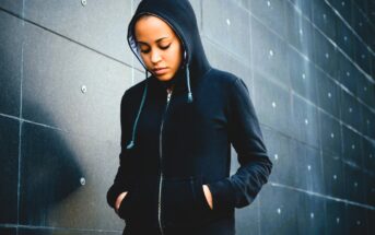 A person wearing a black hoodie stands against a textured, dark gray wall. They are looking downward with hands in the pockets of the hoodie, creating a pensive and introspective mood.