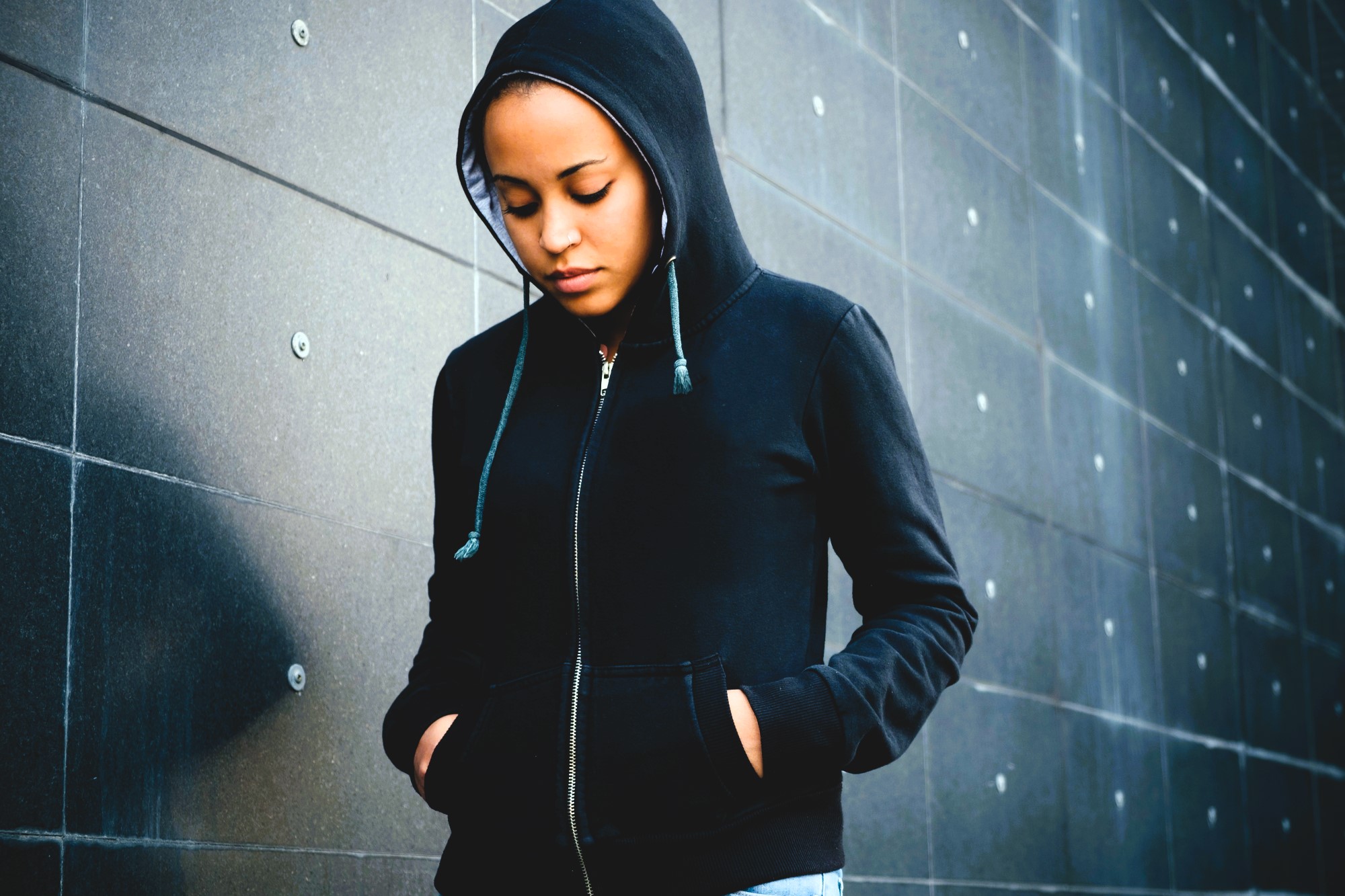 A person wearing a black hoodie stands against a textured, dark gray wall. They are looking downward with hands in the pockets of the hoodie, creating a pensive and introspective mood.