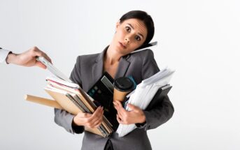 A woman in a gray suit, looking overwhelmed, balances a phone between her ear and shoulder while holding numerous folders, notebooks, a calculator, and a coffee cup. A hand from the side offers more documents.