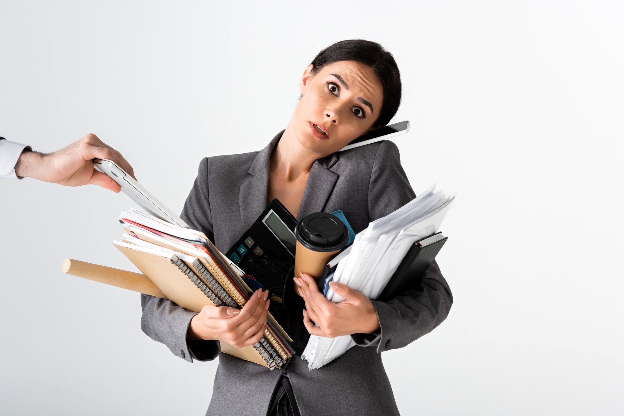 A woman in a gray suit, looking overwhelmed, balances a phone between her ear and shoulder while holding numerous folders, notebooks, a calculator, and a coffee cup. A hand from the side offers more documents.