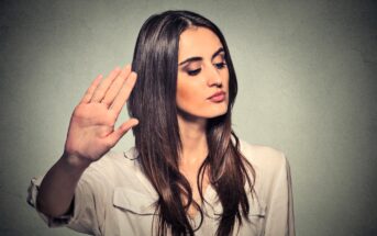 A woman with long brown hair is holding up her hand with her palm facing outward, as if gesturing "stop" or "talk to the hand." She is wearing a white shirt and looking to the side against a textured gray background.