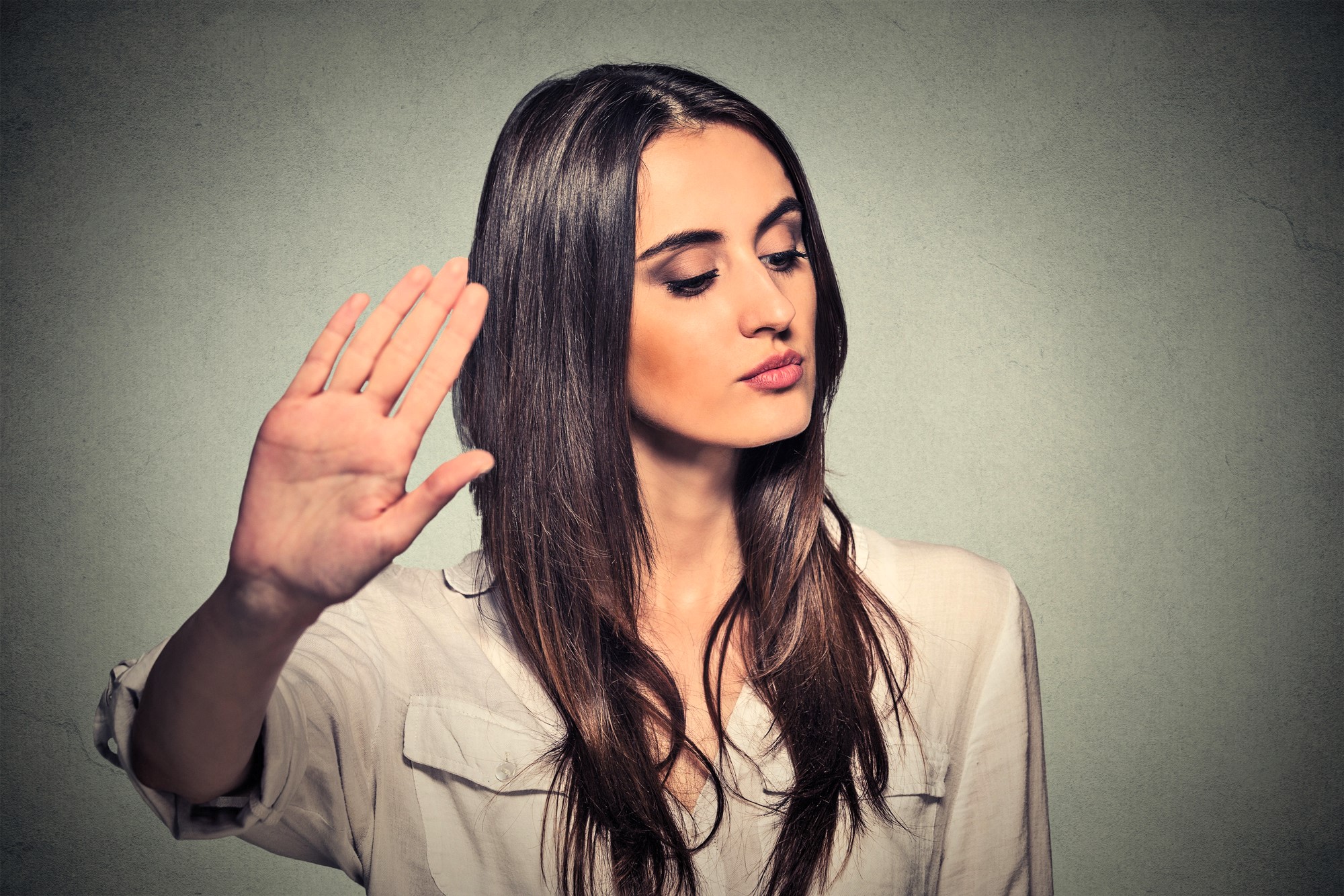 A woman with long brown hair is holding up her hand with her palm facing outward, as if gesturing "stop" or "talk to the hand." She is wearing a white shirt and looking to the side against a textured gray background.