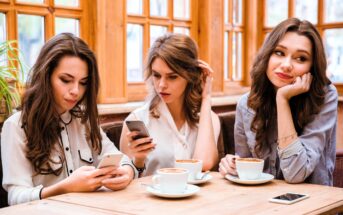 Three women are seated at a wooden table with coffee cups in a cozy cafe. Two are looking at their smartphones, while the third leans her head on her hand, gazing away thoughtfully. Large windows with wooden frames are in the background.