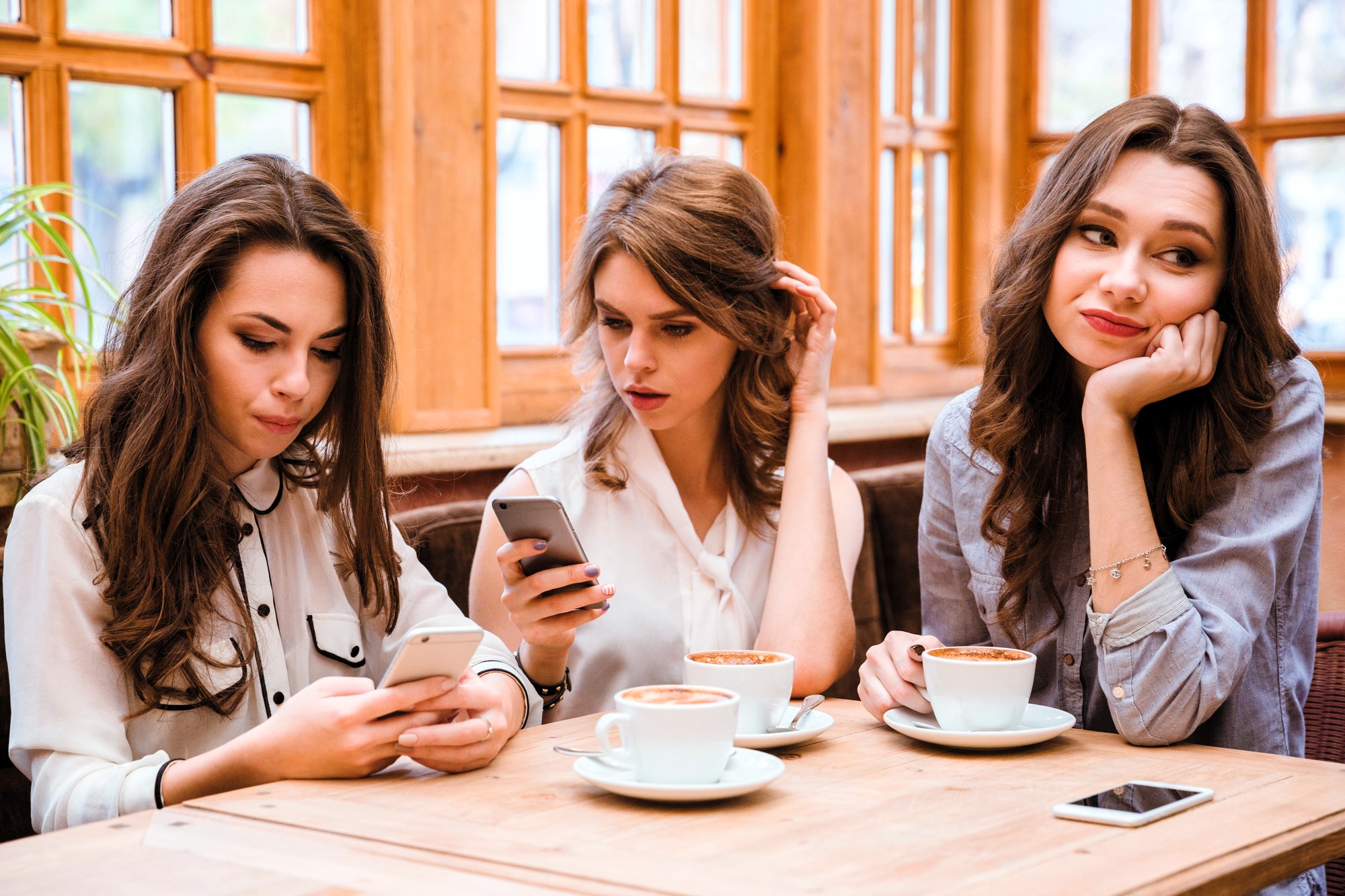 Three women are seated at a wooden table with coffee cups in a cozy cafe. Two are looking at their smartphones, while the third leans her head on her hand, gazing away thoughtfully. Large windows with wooden frames are in the background.