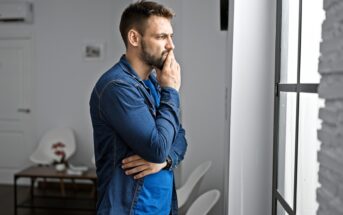 A man with a beard, wearing a denim shirt, stands indoors with his hand on his chin, gazing thoughtfully out of a window. A small table and chairs are in the background.