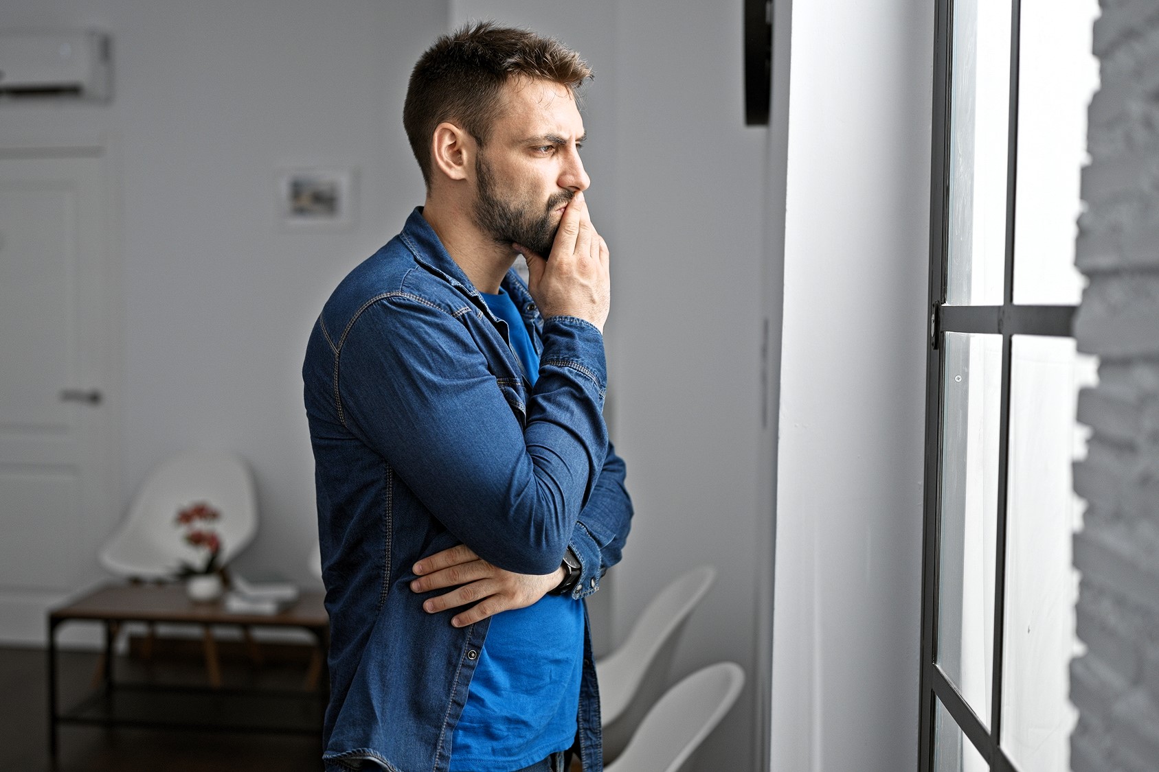 A man with a beard, wearing a denim shirt, stands indoors with his hand on his chin, gazing thoughtfully out of a window. A small table and chairs are in the background.