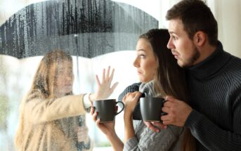 A couple holding mugs looks outside at a woman under an umbrella. The woman appears to be wet from the rain and is pressing a hand against a glass window, looking at the couple inside. The outdoor scene is rainy and the window is covered in raindrops.