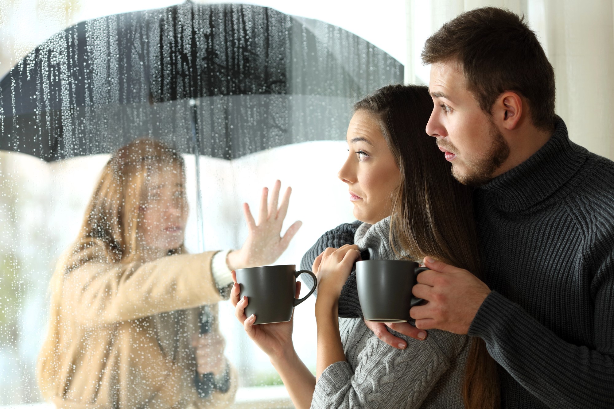 A couple holding mugs looks outside at a woman under an umbrella. The woman appears to be wet from the rain and is pressing a hand against a glass window, looking at the couple inside. The outdoor scene is rainy and the window is covered in raindrops.