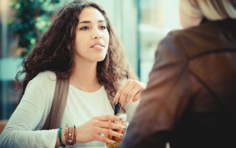 A woman with long curly hair sits at a table, holding a glass with a straw. She wears a white shirt and bracelets, looking thoughtful. Another person is partially visible, facing her. The background is blurred.