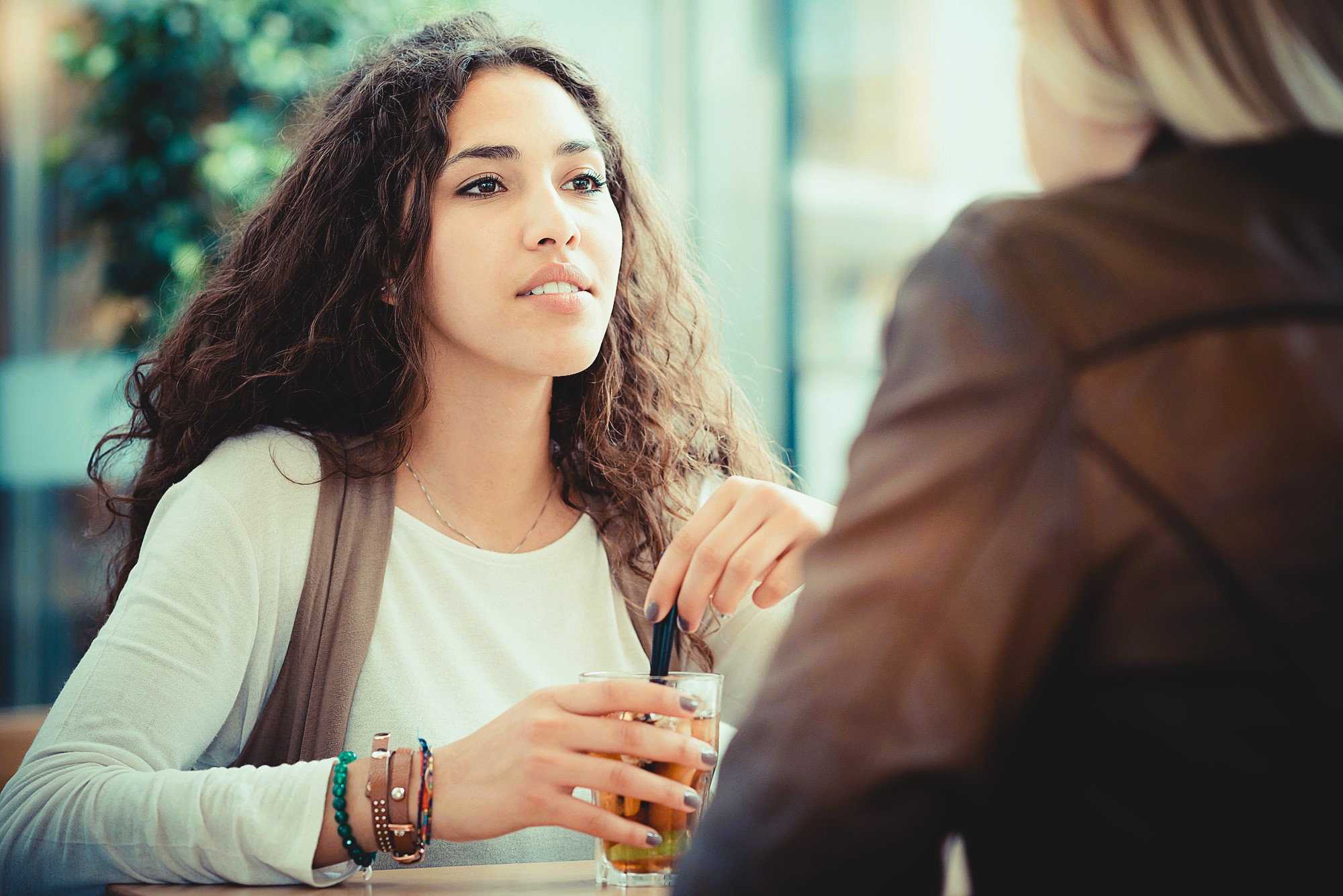 A woman with long curly hair sits at a table, holding a glass with a straw. She wears a white shirt and bracelets, looking thoughtful. Another person is partially visible, facing her. The background is blurred.