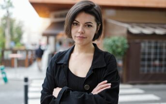 A person with short dark hair wearing a black jacket stands on a city street with arms crossed. They have a neutral expression, and the background shows a blurred view of outdoor seating and buildings.