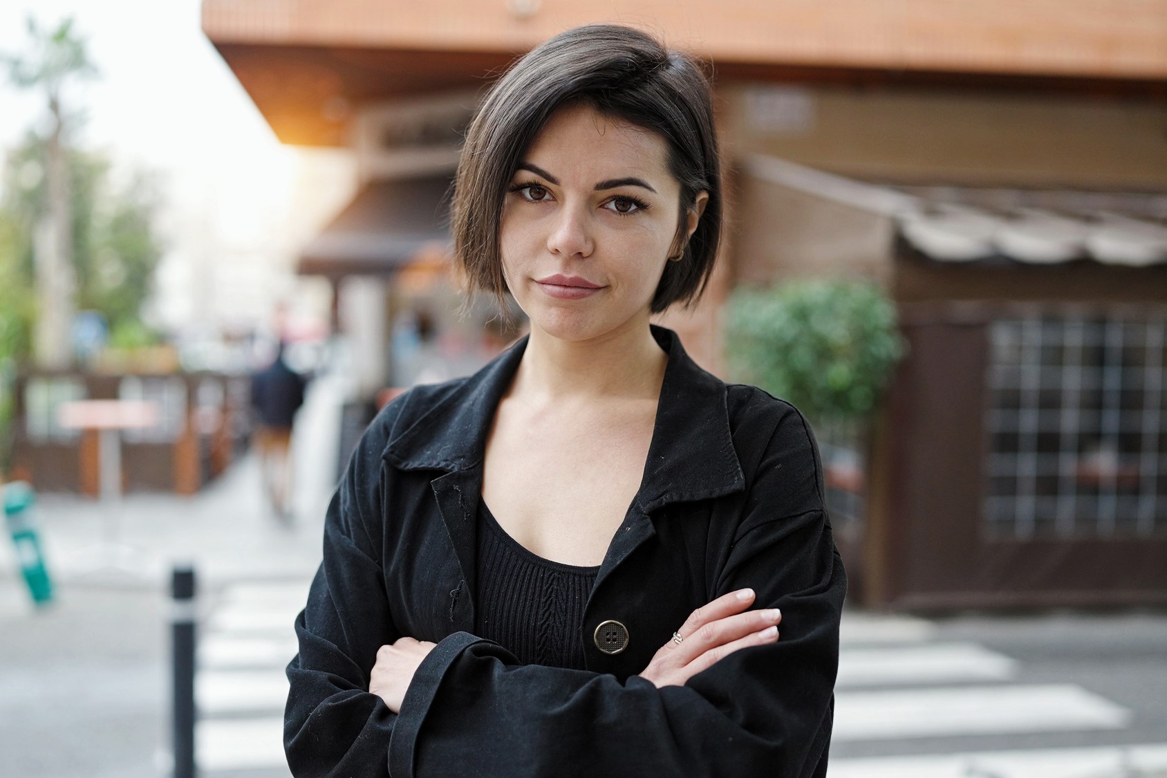 A person with short dark hair wearing a black jacket stands on a city street with arms crossed. They have a neutral expression, and the background shows a blurred view of outdoor seating and buildings.