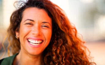 Smiling person with curly hair and a nose piercing, wearing a green shirt, captured in bright lighting. The background is softly blurred, emphasizing their joyful expression.