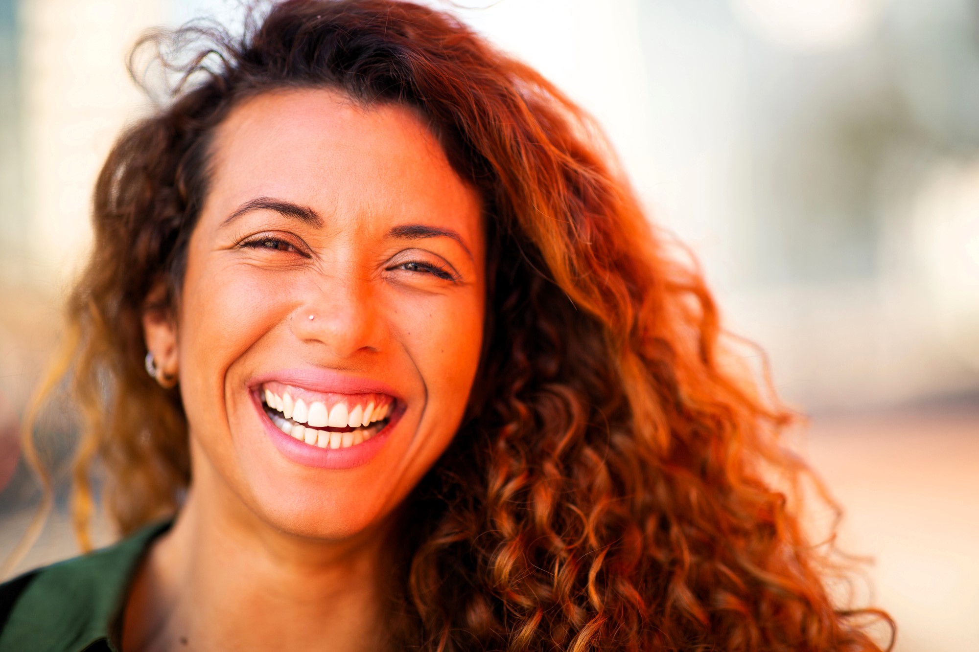 Smiling person with curly hair and a nose piercing, wearing a green shirt, captured in bright lighting. The background is softly blurred, emphasizing their joyful expression.