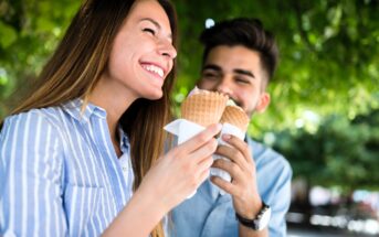 A smiling woman and man enjoy ice cream cones outdoors under green trees. The woman wears a blue striped shirt, and the man has a beard and wears a light blue shirt. They appear happy and relaxed.