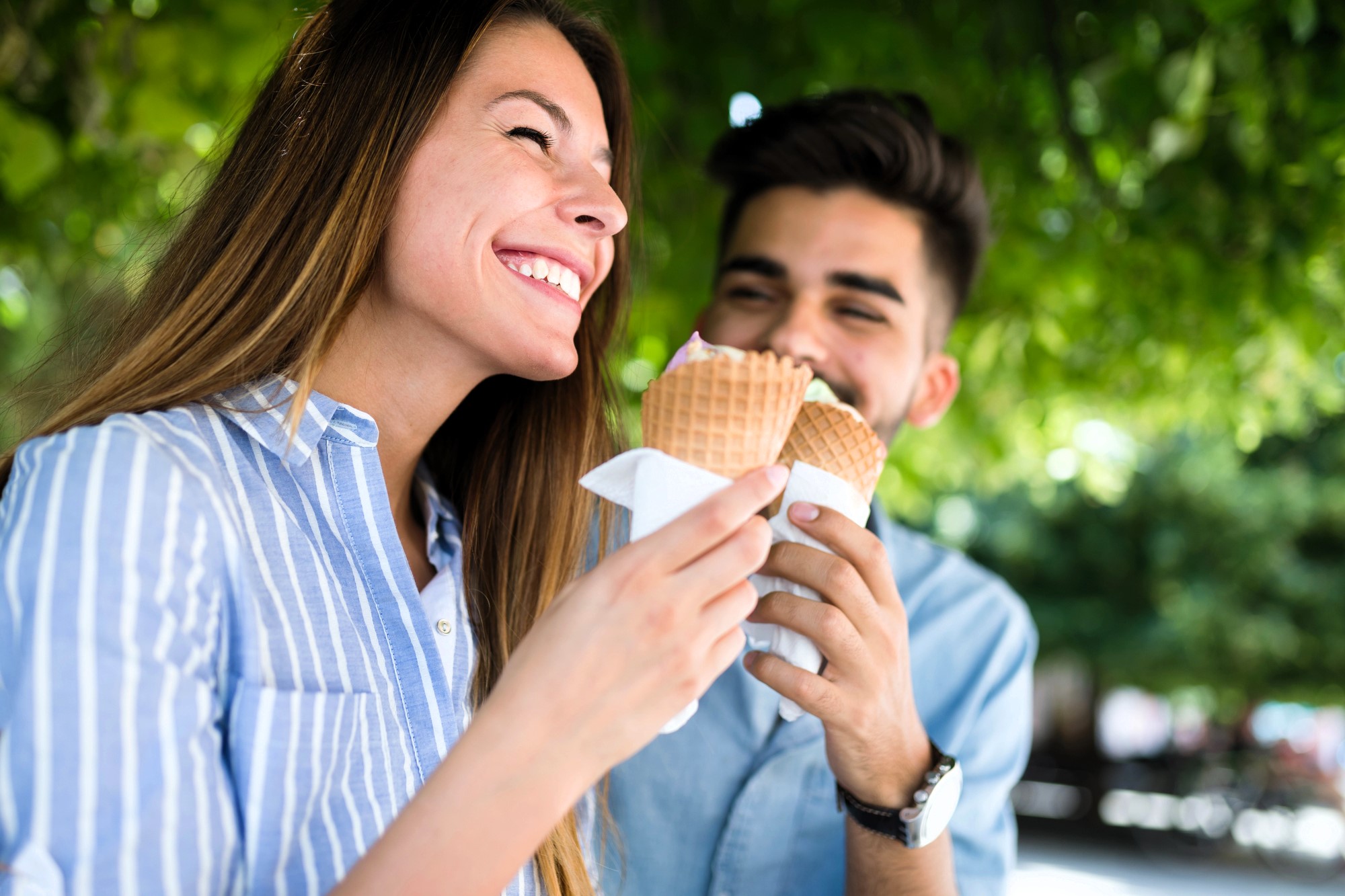 A smiling woman and man enjoy ice cream cones outdoors under green trees. The woman wears a blue striped shirt, and the man has a beard and wears a light blue shirt. They appear happy and relaxed.