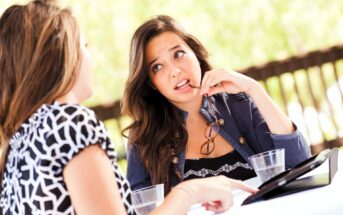 Two women are engaged in a conversation at an outdoor table. One woman, holding glasses to her lips, listens attentively. Sunlight and greenery are in the background, with plastic cups and a tablet on the table.