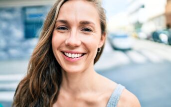 A woman with long hair smiles at the camera, standing outdoors on a sunny day. She is wearing a sleeveless top, and the background shows a blurred urban street scene with buildings and a car.