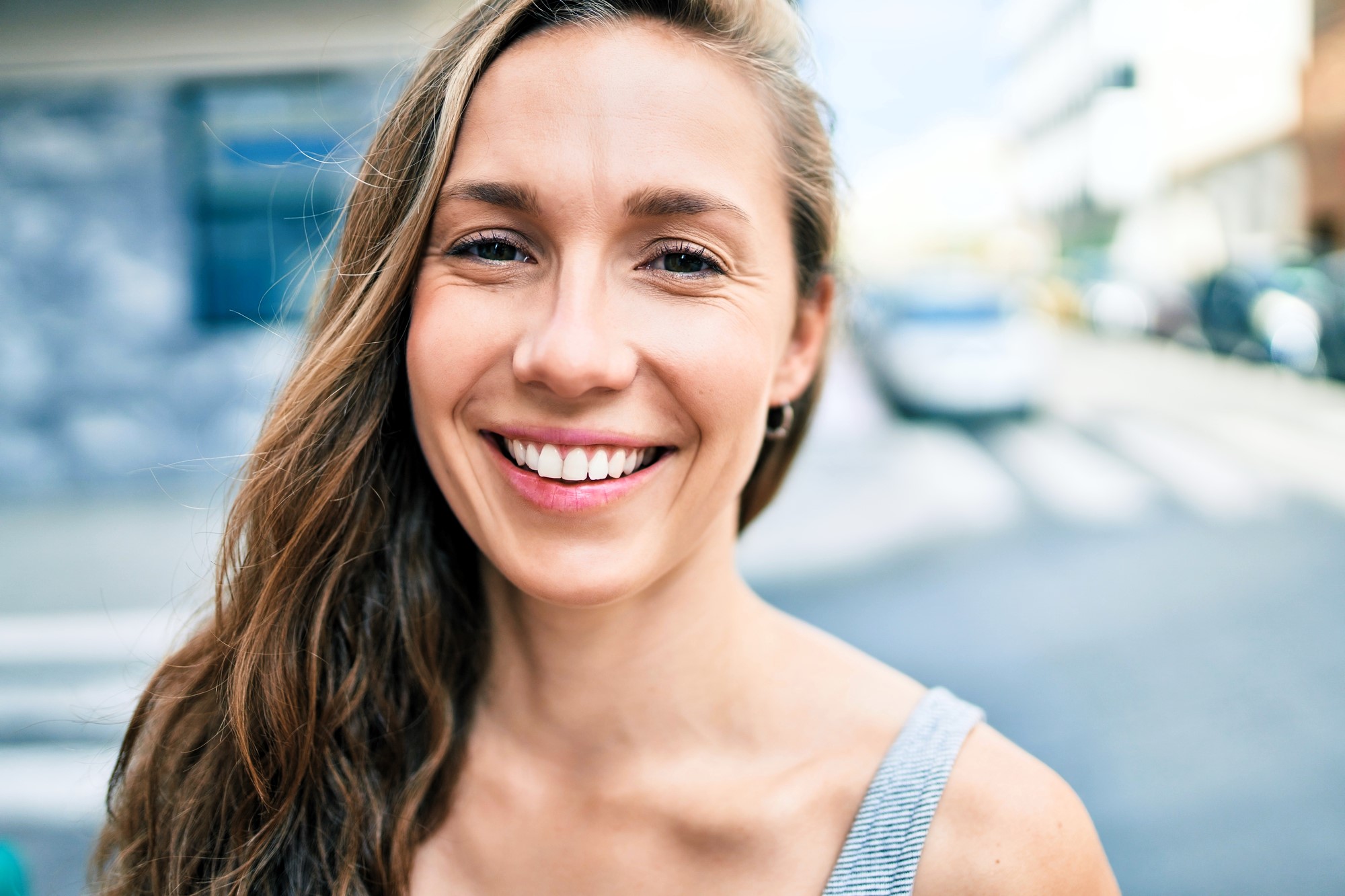 A woman with long hair smiles at the camera, standing outdoors on a sunny day. She is wearing a sleeveless top, and the background shows a blurred urban street scene with buildings and a car.