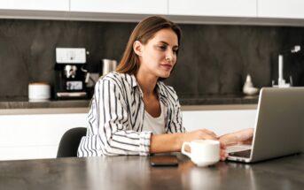 Woman in a striped shirt sits at a kitchen table, using a laptop. She holds a cup of coffee, with a smartphone nearby. The background features a coffee machine and modern kitchen appliances.