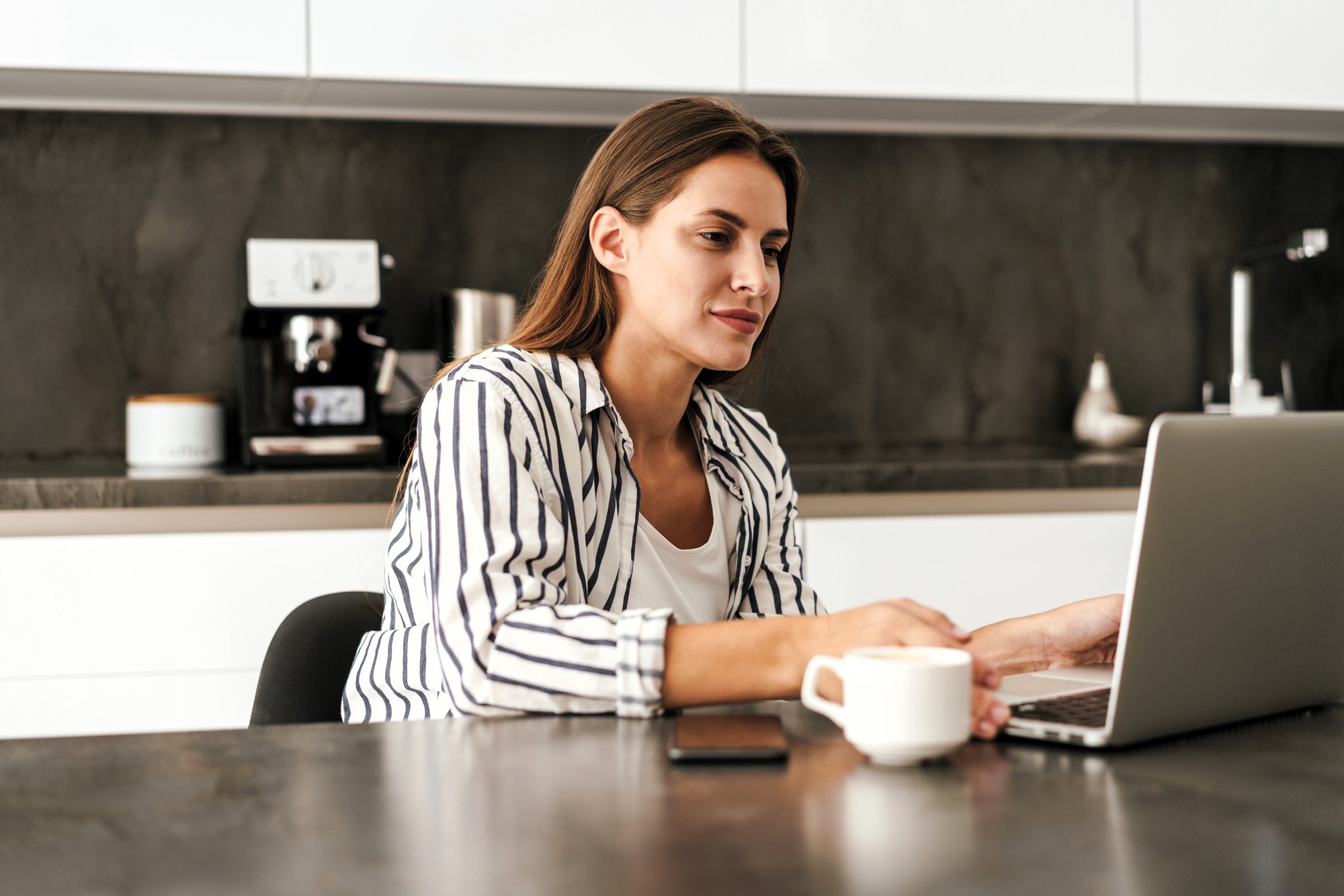 Woman in a striped shirt sits at a kitchen table, using a laptop. She holds a cup of coffee, with a smartphone nearby. The background features a coffee machine and modern kitchen appliances.