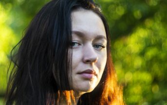 Close-up of a person with long dark hair standing outdoors. They have a neutral expression, and sunlight casts a warm glow on their hair and face. The background is a blurred green, suggesting a natural setting.