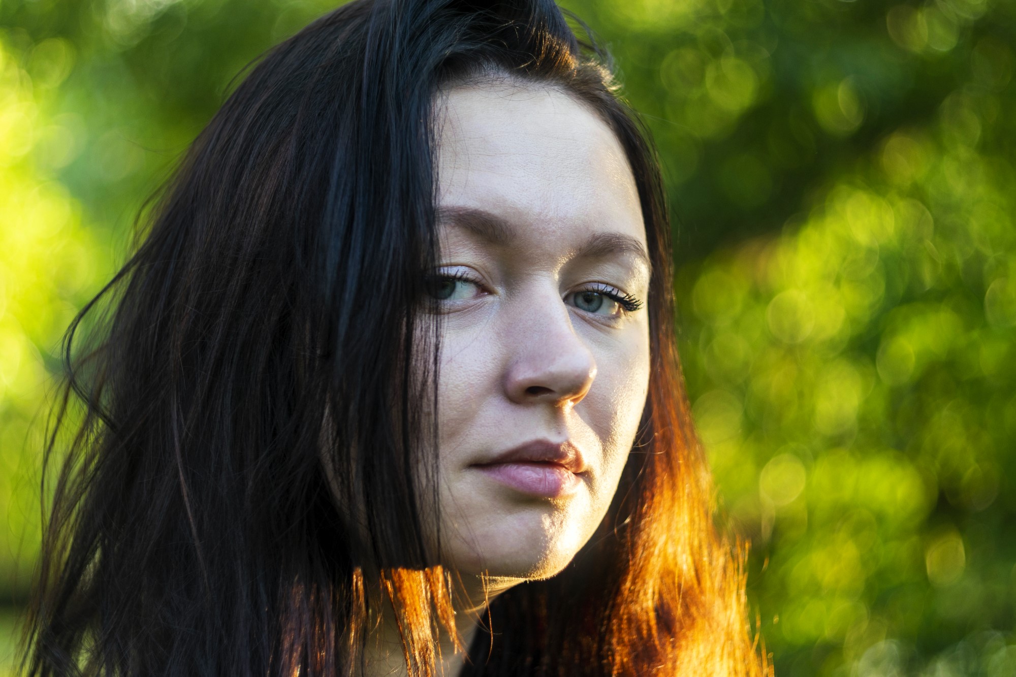 Close-up of a person with long dark hair standing outdoors. They have a neutral expression, and sunlight casts a warm glow on their hair and face. The background is a blurred green, suggesting a natural setting.