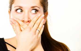 A woman with long brown hair covers her mouth with both hands, her eyes looking to the side. She has a surprised or shocked expression and wears a black top. The background is plain white.