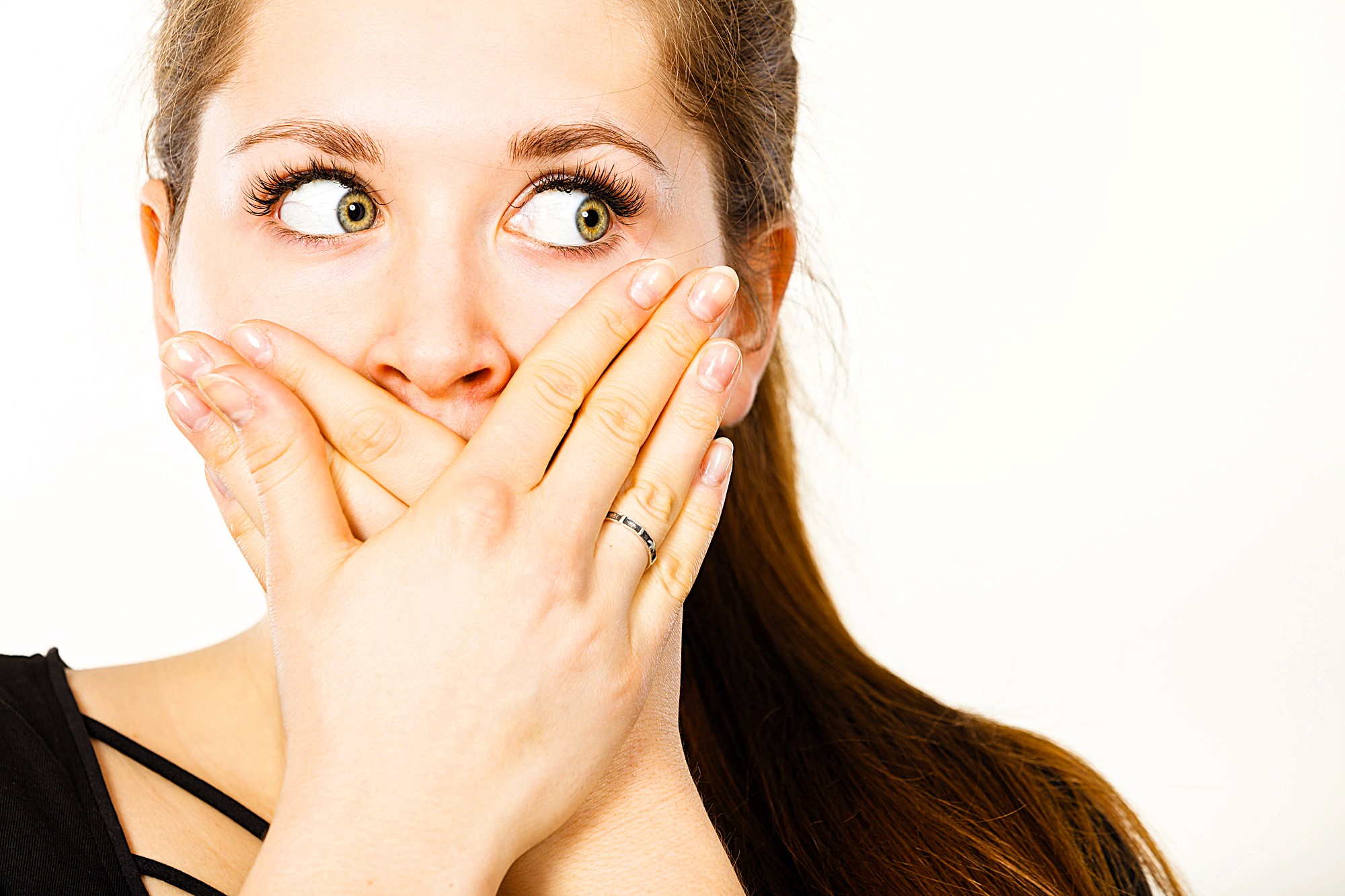 A woman with long brown hair covers her mouth with both hands, her eyes looking to the side. She has a surprised or shocked expression and wears a black top. The background is plain white.