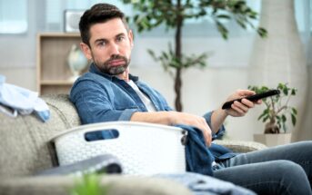 A man sitting on a couch holding a remote control, wearing a denim shirt and jeans. A laundry basket is beside him. A potted plant and some household items can be seen in the background.