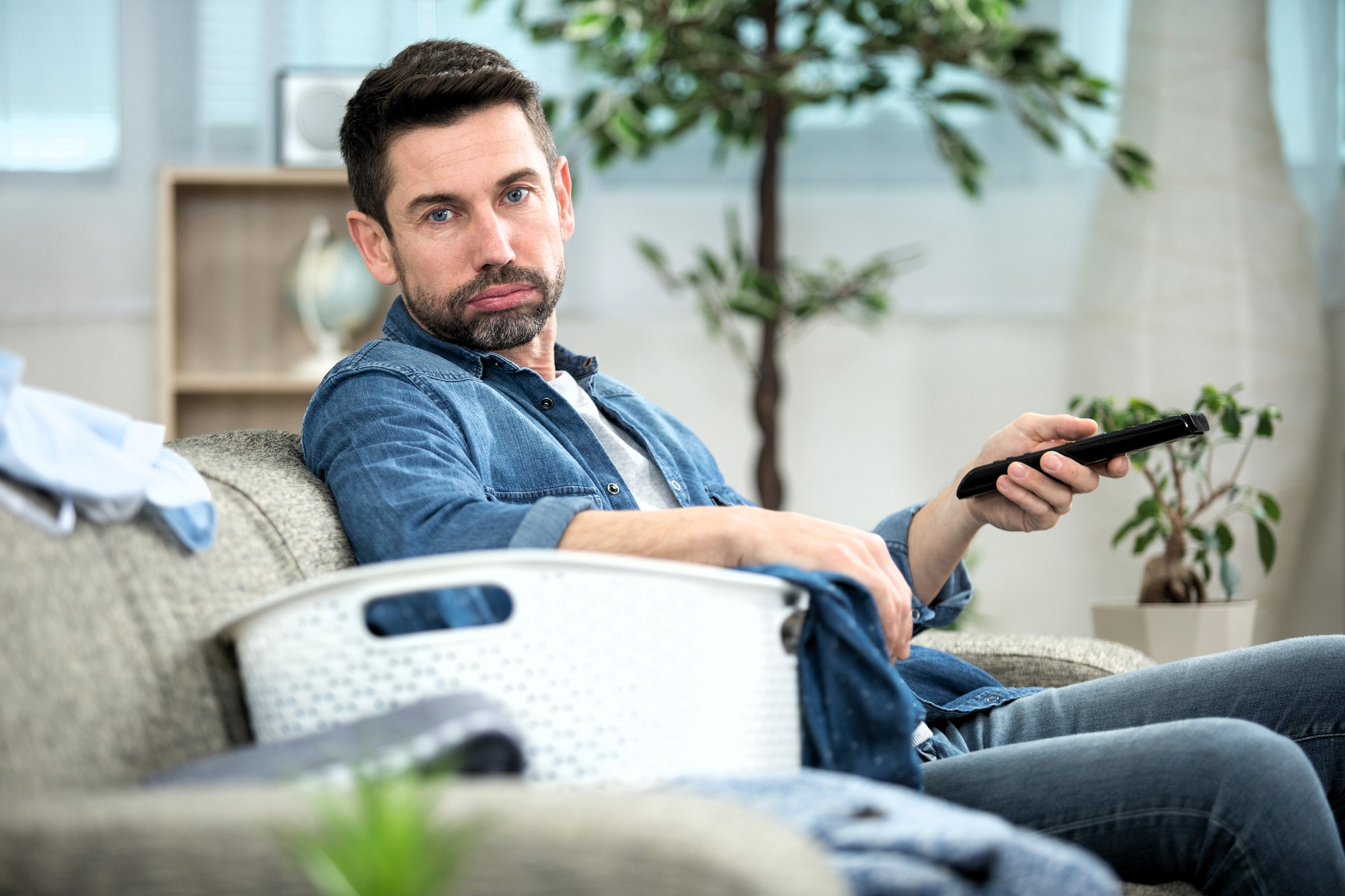 A man sitting on a couch holding a remote control, wearing a denim shirt and jeans. A laundry basket is beside him. A potted plant and some household items can be seen in the background.