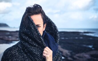 A person is partially obscured by a dark patterned coat on a rocky beach, holding the collar up with one hand. The background shows a cloudy sky and distant water.