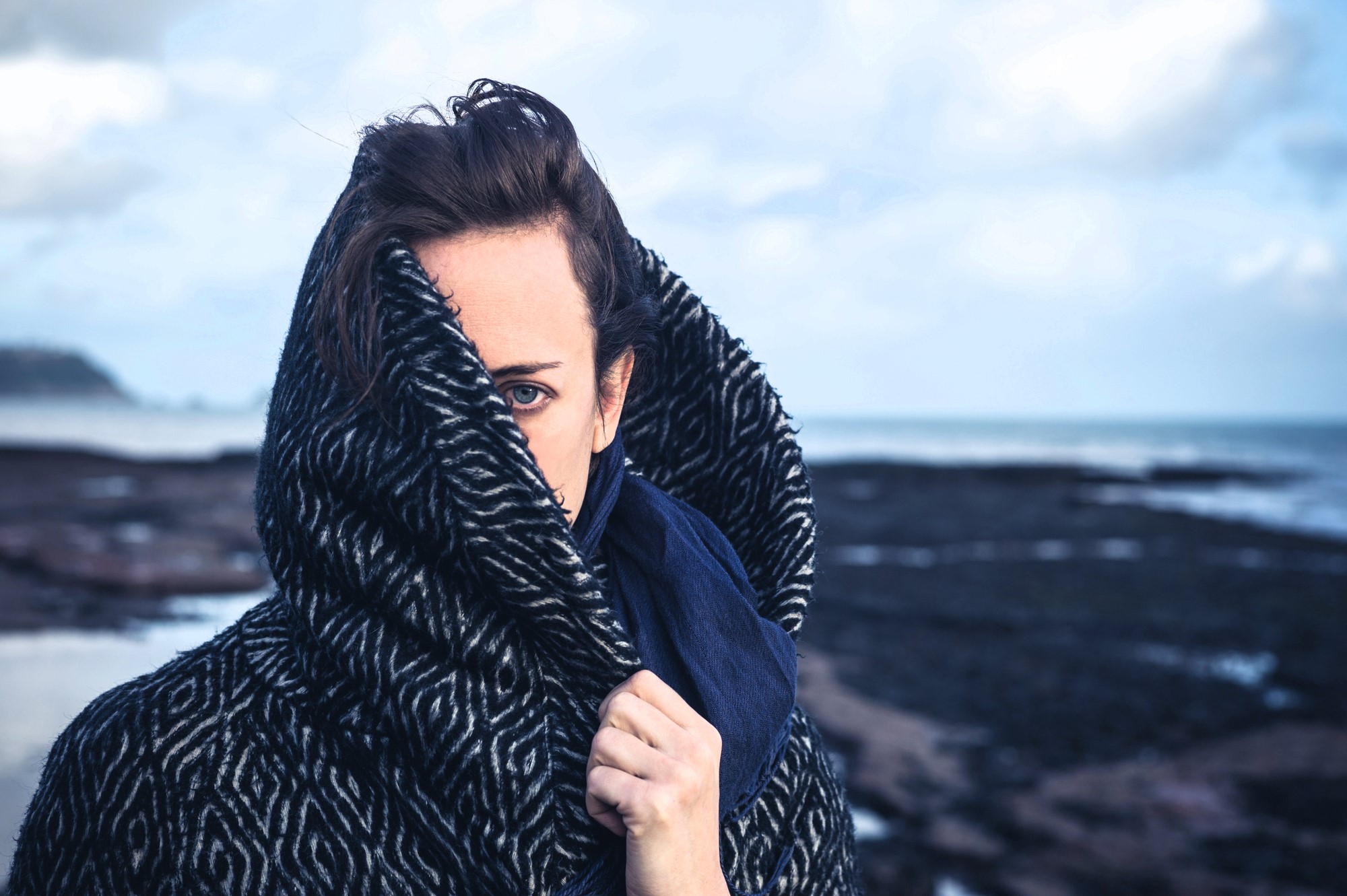 A person is partially obscured by a dark patterned coat on a rocky beach, holding the collar up with one hand. The background shows a cloudy sky and distant water.