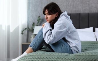 A woman in a gray hoodie and blue jeans sits on a bed with a green knitted blanket, hugging her knees and looking thoughtful. The room is softly lit with a potted plant and a lamp in the background.