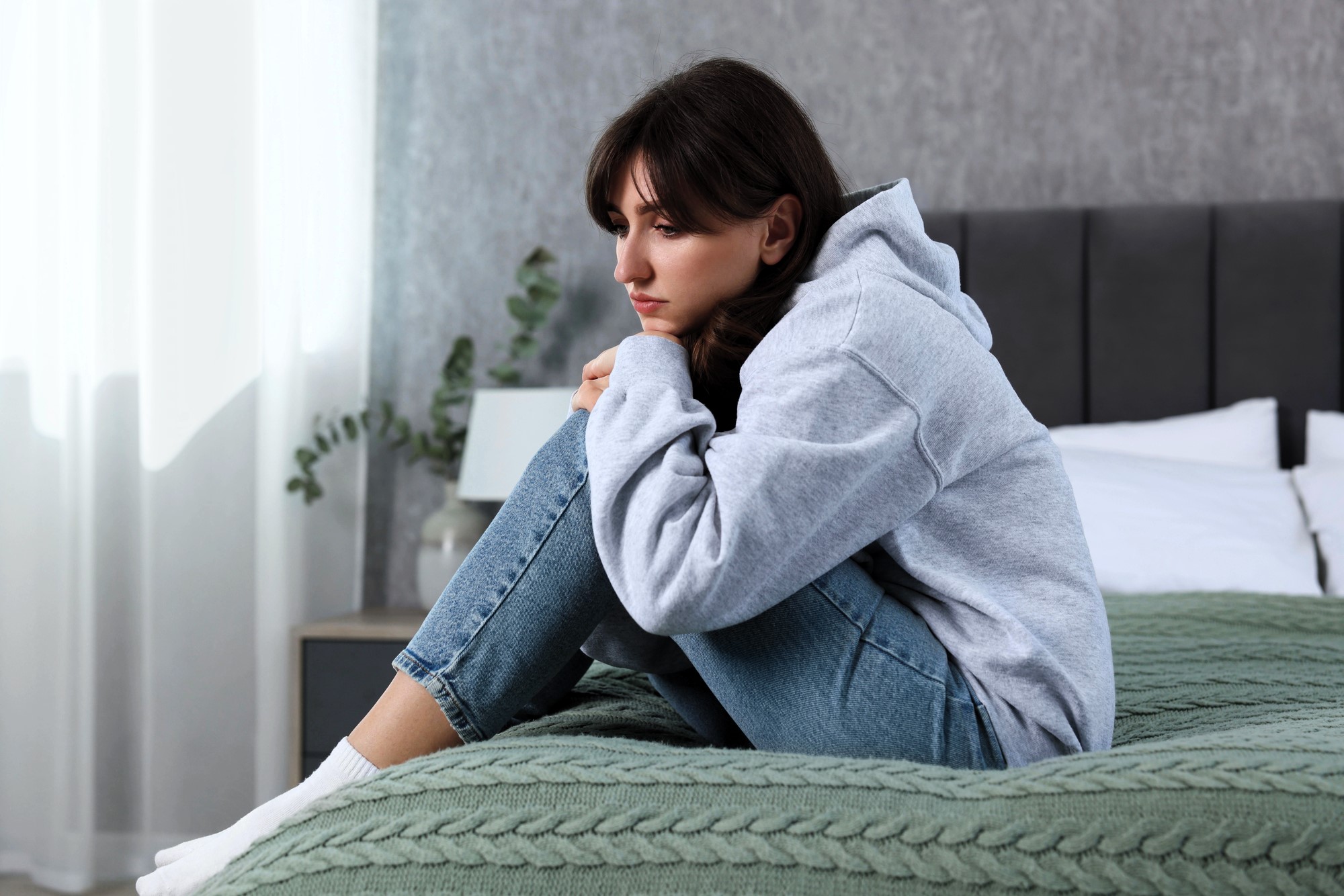 A woman in a gray hoodie and blue jeans sits on a bed with a green knitted blanket, hugging her knees and looking thoughtful. The room is softly lit with a potted plant and a lamp in the background.