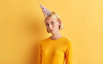 A person with short blonde hair wearing a yellow shirt and a polka-dotted party hat stands against a matching yellow background, looking slightly pensive.