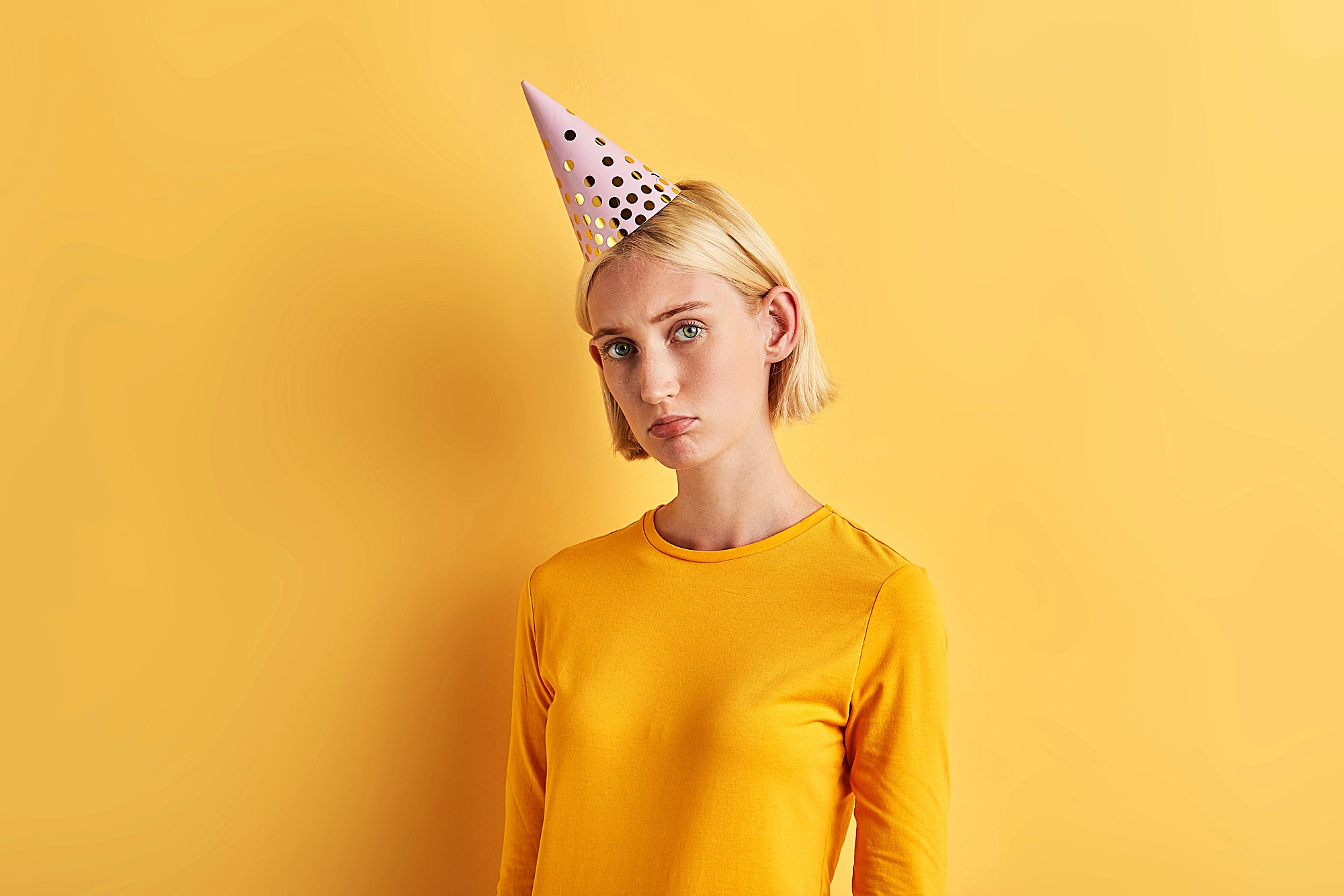 A person with short blonde hair wearing a yellow shirt and a polka-dotted party hat stands against a matching yellow background, looking slightly pensive.