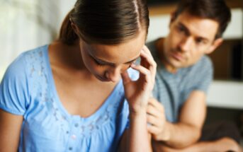 A woman in a blue shirt looks down, appearing upset, while a man in a striped shirt beside her extends a hand in support. The background is softly blurred, focusing attention on their interaction.