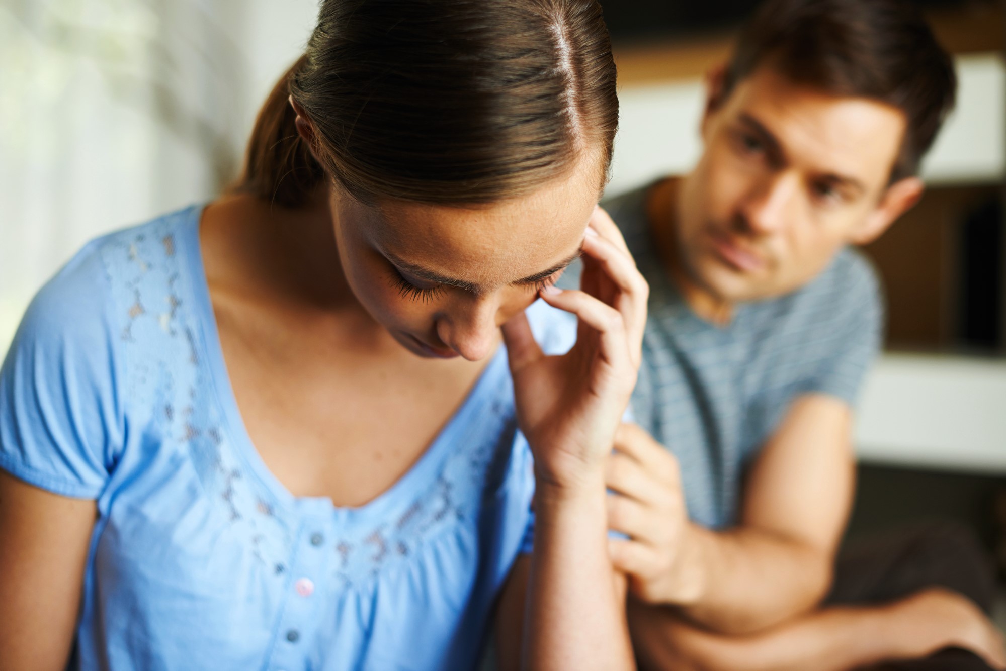 A woman in a blue shirt looks down, appearing upset, while a man in a striped shirt beside her extends a hand in support. The background is softly blurred, focusing attention on their interaction.