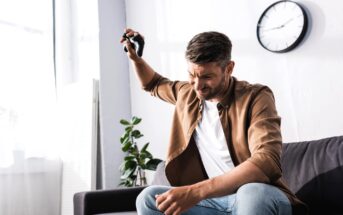 A man sitting on a couch, holding a video game controller above his head in frustration. He is wearing a brown jacket and jeans. A potted plant and clock are in the background.