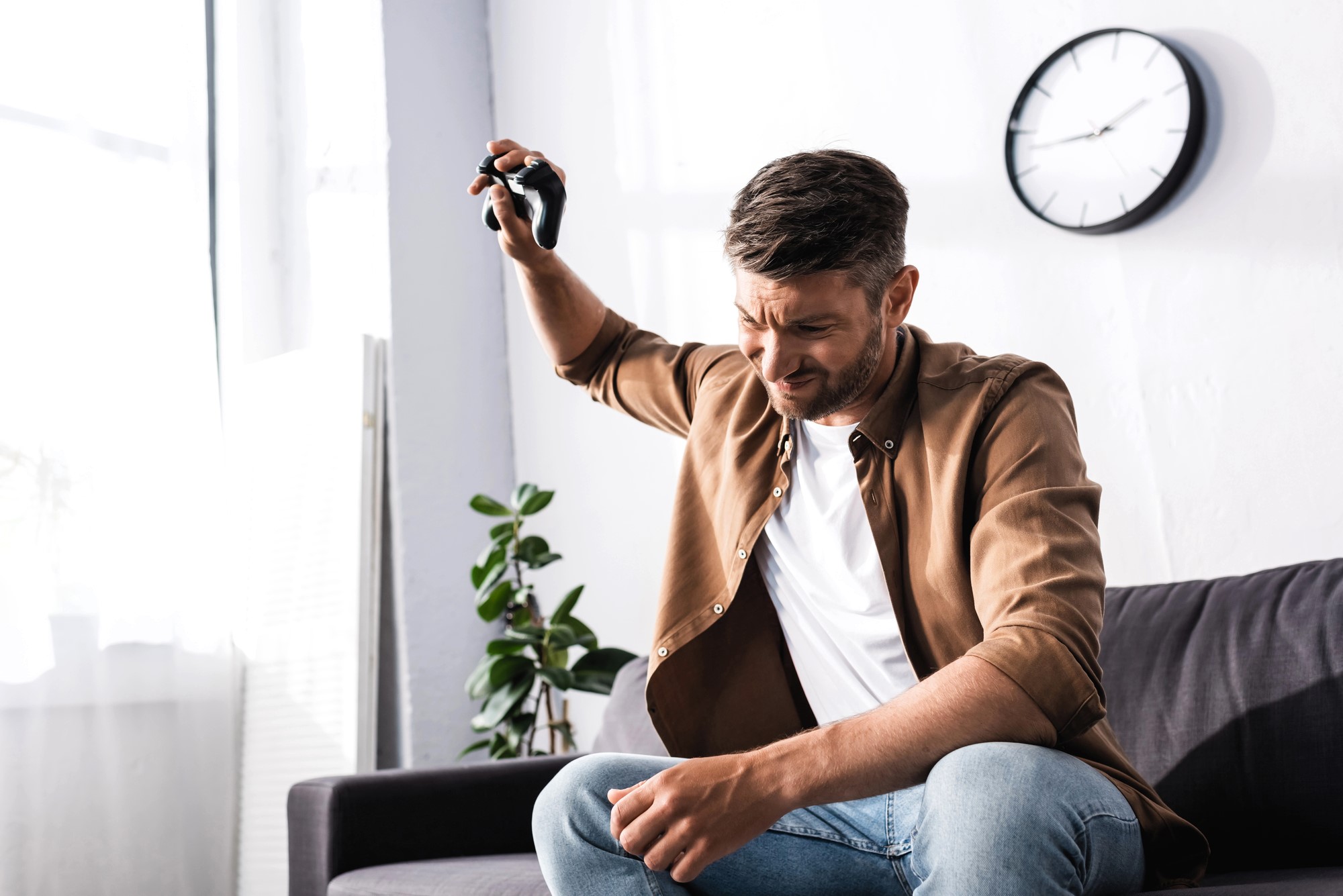 A man sitting on a couch, holding a video game controller above his head in frustration. He is wearing a brown jacket and jeans. A potted plant and clock are in the background.