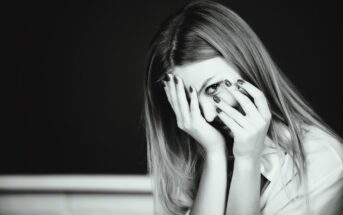 Black and white image of a woman with long hair, partially covering her face with her hands. Her eyes are visible through her fingers, and she has polished nails. She appears thoughtful or pensive, set against a dark background.