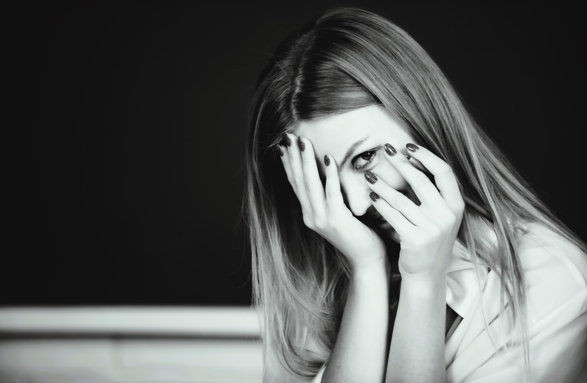 Black and white image of a woman with long hair, partially covering her face with her hands. Her eyes are visible through her fingers, and she has polished nails. She appears thoughtful or pensive, set against a dark background.