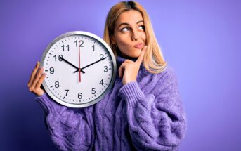 A person with long blonde hair, wearing a purple sweater, holds a large clock showing 8:20. They are looking upward with a thoughtful expression against a purple background.